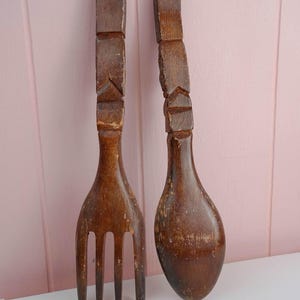 May include: A pair of vintage wooden salad servers. One is a fork with four tines, the other a spoon. Both have carved geometric designs on the handles and are finished in a dark brown stain. The fork measures approximately 12 inches long.