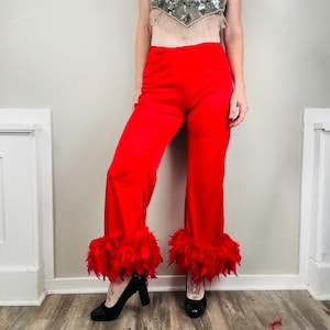May include: Bright red trousers featuring a high-waisted fit and wide legs that end in a thick, matching red feather trim at the ankles. The pants are paired with a sleeveless, cropped halter top adorned with silver sequins and beads.