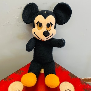 May include: A vintage plush toy of a cartoon mouse character. The toy has a black body, large black ears, and a cream-colored face with large eyes. The mouse has yellow feet and is standing on a red surface.