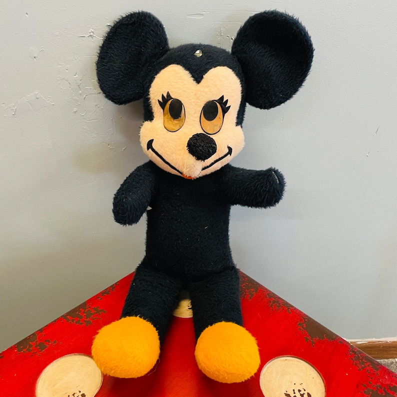 May include: A vintage plush toy of a cartoon mouse character. The toy has a black body, large black ears, and a cream-colored face with large eyes. The mouse has yellow feet and is standing on a red surface.