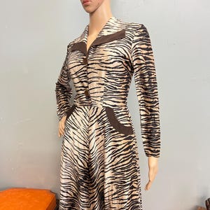 May include: A long-sleeved dress featuring a tan and black tiger stripe pattern. The garment includes a solid brown pointed collar, two brown buttons down the front bodice, and two brown curved pocket accents at the waistline of the skirt.