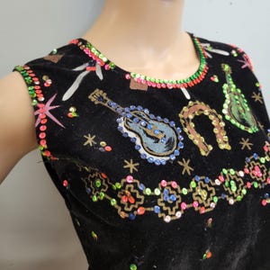 May include: A black sleeveless top featuring colorful sequin trim around the neckline and armholes. The fabric is decorated with embroidered motifs including guitars, a horseshoe, and stars, accented with scattered sequins and geometric patterns.