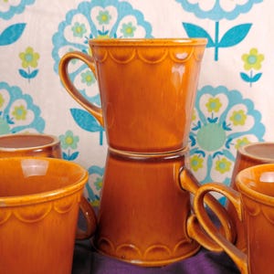 May include: A collection of orange ceramic mugs with scalloped rims and curved handles. The mugs are arranged in a group, some stacked, against a backdrop of a floral patterned fabric in shades of blue, green, and white. The mugs have a glossy finish.