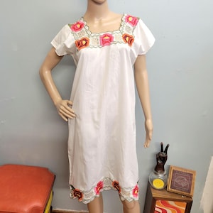 May include: A white knee-length dress featuring a square neckline and short sleeves. The garment is decorated with colorful floral embroidery in shades of pink, orange, and green around the neckline and along the scalloped bottom hem.