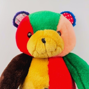 May include: A plush teddy bear constructed from multi-colored fabric patches. The bear features a bright yellow muzzle, a red and green head, a brown arm, and a green arm. The ears are accented with purple, plaid, and red-and-white polka dot patterns.