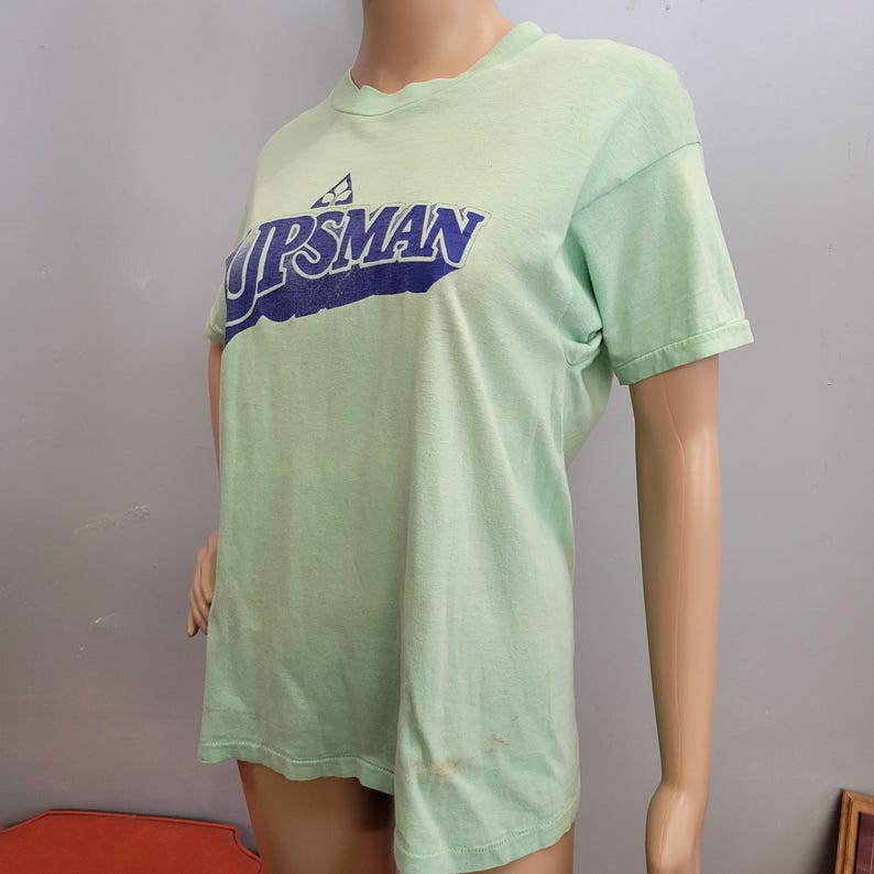 May include: A light green t-shirt with the word "UPSMAN" in blue lettering. The shirt has short sleeves and a crew neck. The shirt is displayed on a mannequin.