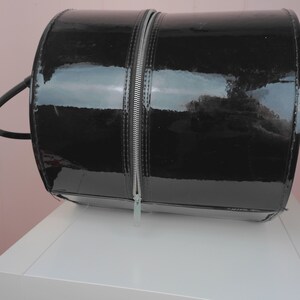 May include: A black, cylindrical handbag with a glossy patent leather finish. The bag features a full-length silver zipper down the center and a small handle on the side. The bag is sitting on a white surface.