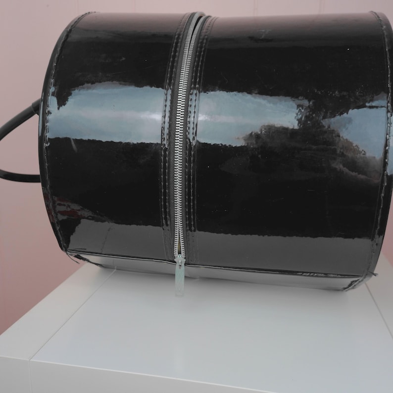May include: A black, cylindrical handbag with a glossy patent leather finish. The bag features a full-length silver zipper down the center and a small handle on the side. The bag is sitting on a white surface.