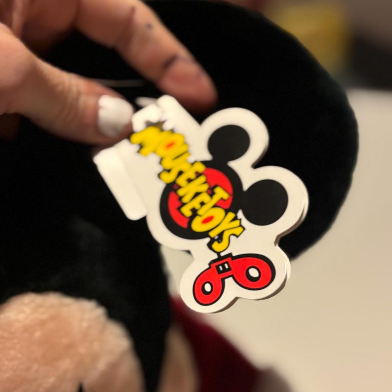Pode incluir: Um brinquedo de pel&uacute;cia preto com uma etiqueta vermelha e branca que diz "MOUSEKETOYS" em amarelo e vermelho. A etiqueta tem a forma da cabe&ccedil;a do Mickey Mouse. O brinquedo tem uma gravata borboleta vermelha.