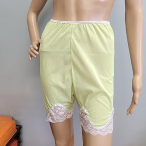 Puede incluir: Pantalones cortos de color verde lima claro con una cintura elástica blanca y un borde de encaje floral blanco a lo largo de la parte inferior de cada pierna. La prenda está hecha de una tela suave y ligera que llega hasta la mitad del muslo.