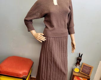 May include: A brown knit sweater set with a collared neckline and a pleated skirt. The sweater has a white collar and a textured pattern on the upper chest. The skirt is a midi length. The set is displayed on a mannequin.
