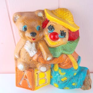 May include: A painted wall plaque featuring a clown with a yellow hat, red nose, and green ruffled collar hugging a brown teddy bear. The bear sits atop a large orange and yellow block marked with the letters A and B. The clown wears blue overalls.