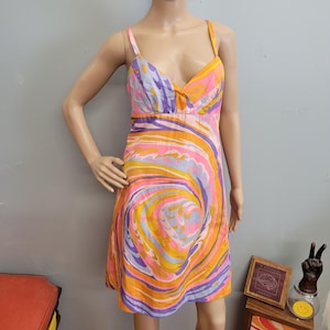 May include: A colorful, vintage-style dress with a swirling pattern in shades of orange, pink, purple, and yellow. The dress features a V-neckline, spaghetti straps, and a fitted bodice that flows into a knee-length skirt. The dress is displayed on a mannequin.