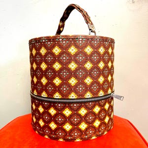 Könnte beinhalten: Eine zylindrische braune Tasche mit einem Blumenmuster in Gelb, Weiß und Grün. Die Tasche hat einen Reißverschluss in der Mitte und einen gemusterten Griff. Die Tasche steht auf einer roten Oberfläche.
