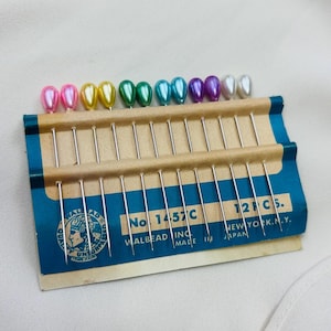 May include: A vintage card of sewing pins with colorful pearlescent heads. The pins are arranged in a row, with colors including pink, yellow, green, and purple. The card has blue and brown accents with text that reads "WALBEAD INC. MADE IN JAPAN."