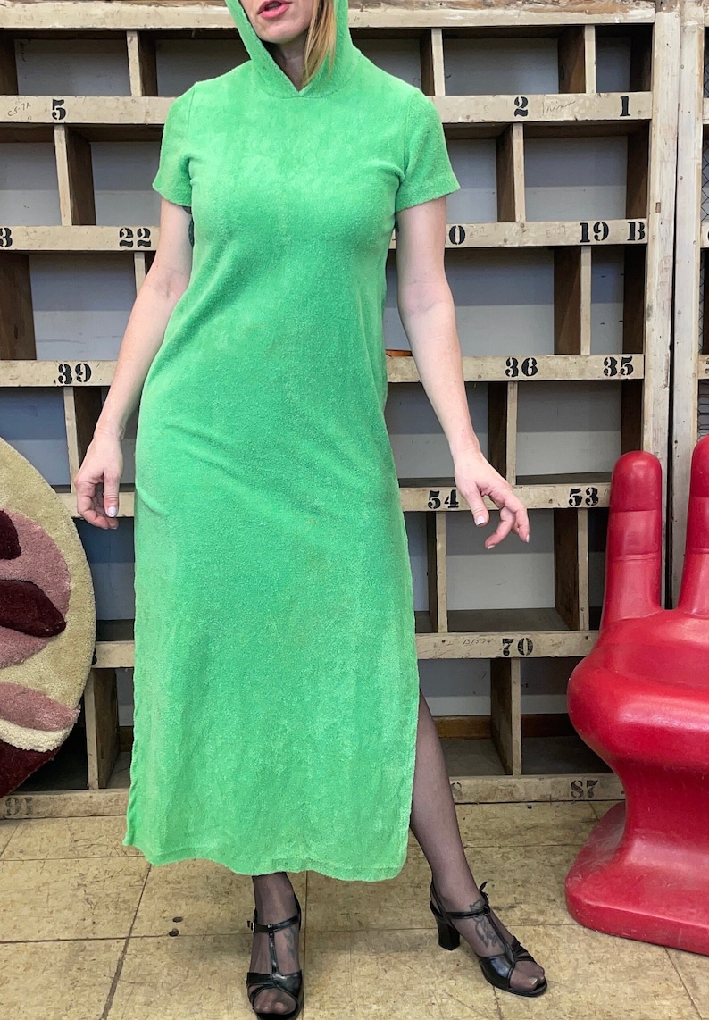 Puede incluir: Un vestido largo verde con capucha y una abertura lateral. El vestido est&aacute; hecho de un tejido suave de felpa. La modelo lleva tacones negros.