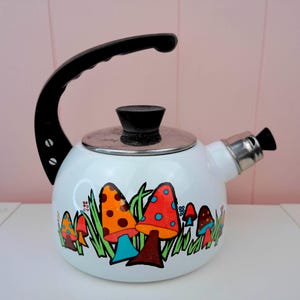 May include: White enamel tea kettle with a black handle and spout. The kettle features a colorful mushroom design with orange, red, and blue caps. The kettle has a silver lid and a black knob on top.
