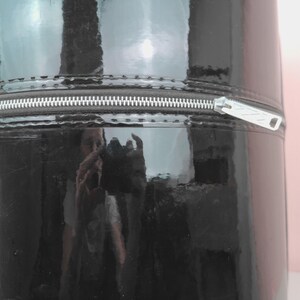 May include: A cylindrical, black, glossy container with a silver zipper. The container's surface reflects the surrounding environment. The zipper runs horizontally across the middle of the container.