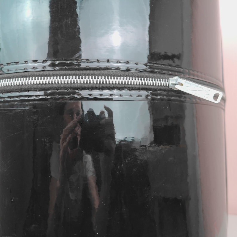 May include: A cylindrical, black, glossy container with a silver zipper. The container's surface reflects the surrounding environment. The zipper runs horizontally across the middle of the container.
