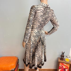 May include: A long-sleeved dress featuring a black and beige tiger stripe pattern. The garment has a high neckline, a fitted bodice, and a flared skirt that falls below the knee. The hem of the dress is finished with a solid black border.