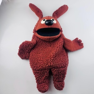 May include: A rust-colored plush puppet featuring a textured, curly fabric body, two long upright ears, and a wide open mouth. The puppet has a smooth black nose and two white circular eyes with black pupils, giving it a distinct character appearance.