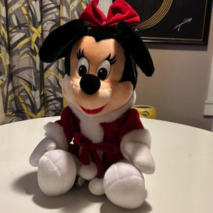 Pode incluir: Uma boneca de pel&uacute;cia da Minnie Mouse vestindo uma roupa de Natal vermelha e branca. A boneca tem orelhas pretas, um la&ccedil;o vermelho e um casaco vermelho e branco. A boneca est&aacute; sentada com luvas e sapatos brancos.