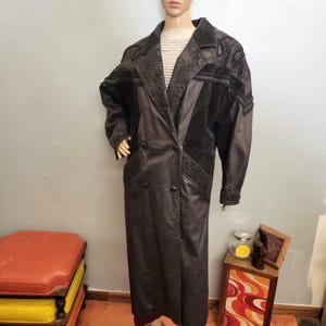 May include: A long, black leather coat with a double-breasted design. The coat features decorative panels with a floral pattern on the shoulders and lapels. The coat is displayed on a mannequin.