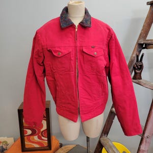 May include: A bright red zip-up jacket featuring a dark, multi-colored patterned collar, long sleeves, and two chest pockets with button closures. The jacket has a central metal zipper and a small black tag on the upper left chest pocket.