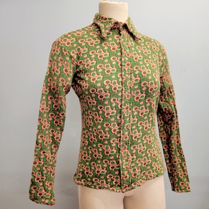 Puede incluir: Camisa abotonada de manga larga con un estampado floral verde y rosa. La camisa tiene un cuello puntiagudo y se exhibe en un maniquí. La tela parece ser un tejido de punto texturizado.