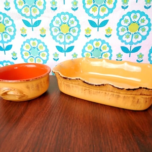 May include: Two vintage ceramic serving dishes. One is a round bowl with an orange interior and yellow exterior with handles. The other is a rectangular baking dish with a scalloped edge and yellow exterior. Both have a distressed look.