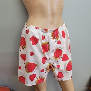 Puede incluir: Pantalones cortos tipo bóxer blancos con un estampado de corazones rojos y detalles dorados. Los pantalones cortos tienen cintura elástica y un corte holgado. El diseño incluye corazones de varios tamaños y elementos decorativos.