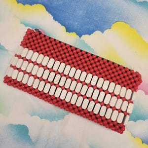 Könnte beinhalten: Eine rechteckige Perlen-Clutch mit Reißverschluss. Die Clutch hat ein Muster aus roten Perlen und weißen rechteckigen Perlen. Der Hintergrund ist ein farbenfrohes, wolkenartiges Muster.