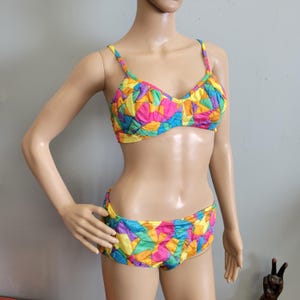 May include: A two-piece bikini set featuring a vibrant, multi-colored geometric patchwork pattern in shades of pink, yellow, blue, and green. The top has thin shoulder straps and a classic cup design, while the matching bottoms offer full coverage.