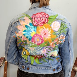 May include: Light blue denim jacket with a vibrant floral design on the back. The hand-painted flowers feature shades of pink, yellow, blue, and green. The jacket has a classic button-up design with two chest pockets and two side pockets.