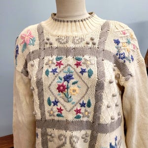 Puede incluir: Suéter color crema con bordado floral. El suéter presenta un cuello alto y mangas largas. El diseño incluye un patrón floral central en forma de diamante con flores rosas, azules y amarillas, rodeado de detalles grises y texturizados.