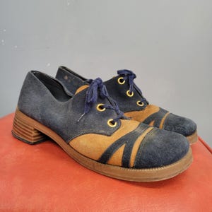 May include: A pair of vintage navy blue and tan suede shoes with a block heel. The shoes have navy blue laces, gold eyelets, and a tan accent design on the toe. The heel measures approximately 2 inches.