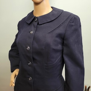 Puede incluir: Una blazer azul marino con cuello redondo y cuatro botones a juego. La blazer tiene un corte entallado y un sutil detalle de diseño en el hombro. Las mangas son largas y la tela parece ser de peso medio.