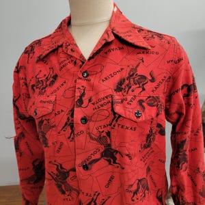 May include: Red button-up shirt with a black print featuring cowboys, horses, and state outlines. The shirt has two chest pockets and black buttons. The print includes the names of states like Arizona, Texas, and Montana.