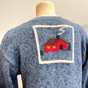 Könnte beinhalten: Blauer Strickpullover mit einem quadratischen, gestickten Aufnäher mit einem roten Haus mit braunem Dach, weißem Rauch und grünen Bäumen. Das Haus hat gelbe Fenster und ist von einem weißen Rahmen umgeben.