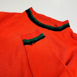 May include: Close-up of an orange sweater with a crew neck and long sleeves. The collar and cuffs feature a black stripe between two green stripes. The sweater is made of a soft, knitted material.