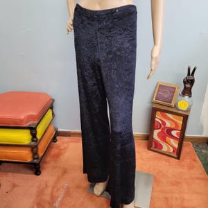 Puede incluir: Pantalones de terciopelo azul marino oscuro con un diseño de pernera ancha. Los pantalones tienen un aspecto de terciopelo arrugado y texturizado y se exhiben en un maniquí. Los pantalones son de color sólido y de cintura alta.