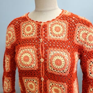 May include: A vintage, button-down crochet cardigan in shades of orange, peach, and cream. The sweater features a square, floral-like pattern throughout, with a scalloped neckline and long sleeves. The buttons are a light brown color.
