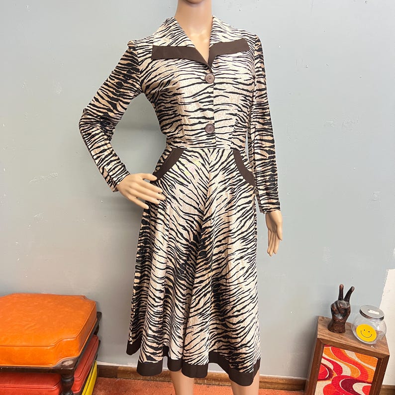 May include: A long-sleeved midi dress featuring a tan and black zebra print pattern. The dress has a pointed collar with dark brown trim, two dark brown buttons on the bodice, dark brown pocket accents at the waist, and a dark brown hemline.