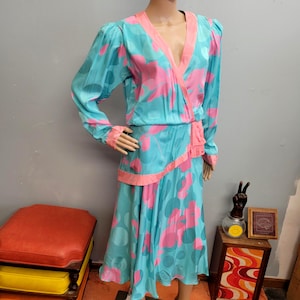 May include: A vintage two-piece dress in a teal and pink floral pattern. The long-sleeved top has a wrap-style front with pink trim, and the skirt is midi-length. The dress is displayed on a mannequin.