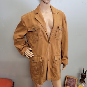 May include: A tan-colored jacket with four front pockets and button closures. The jacket has a collared neckline and long sleeves. The garment is displayed on a mannequin.