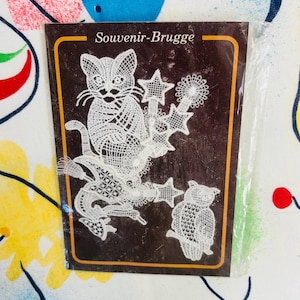 Könnte beinhalten: Eine weiße Spitzenapplikation mit einer Katze, einer Eule und Sternformen auf einer dunkelbraunen rechteckigen Trägerkarte. Der Text Souvenir-Brugge ist oben auf der Karte in Weiß gedruckt, die in einer transparenten Kunststoffhülle steckt.