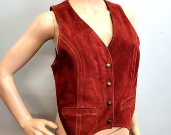 May include: A rust-colored suede vest with a V-neck and four brass-colored buttons. The vest has two small pockets and decorative stitching. This vintage vest is a stylish addition to any wardrobe.