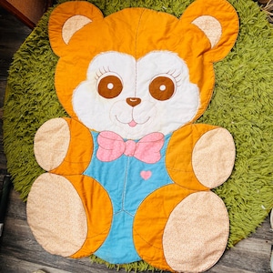 May include: A quilted fabric mat shaped like a teddy bear with orange fur, a white face, and brown eyes. The bear wears a blue shirt with a pink polka-dot bow tie and a small pink heart detail. The ears, hands, and feet feature a light tan floral pattern.