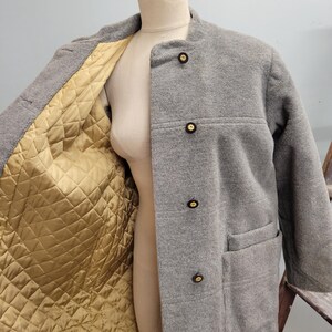 May include: A gray wool coat with a mandarin collar and four dark buttons. The coat has two front pockets and a quilted gold lining. The coat is displayed on a mannequin.