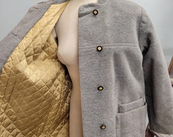 Peut inclure: Un manteau en laine gris avec un col officier et quatre boutons foncés. Le manteau a deux poches avant et une doublure dorée matelassée. Le manteau est présenté sur un mannequin.
